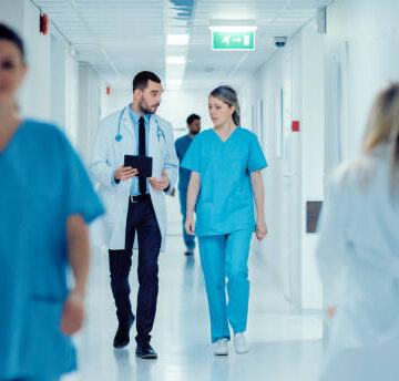 Surgeon and Female Doctor Walk Through Hospital Hallway, They Consult Digital Tablet Computer while Talking about Patient's Health. Modern Bright Hospital with Professional Staff.